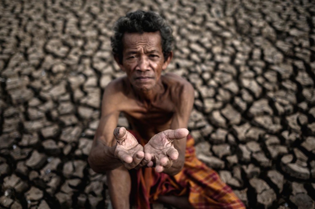 An elderly man on parched, cracked earth holding out cupped hands — a plea for water and relief during drought.